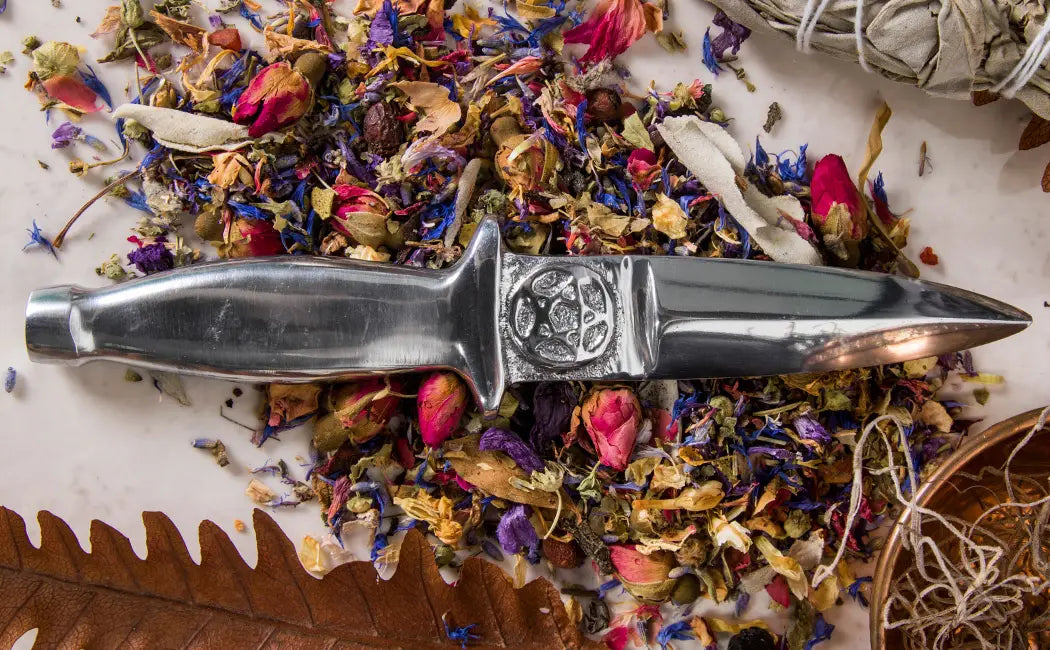 an image of an aluminium athame with a pentacle on the blade. the white background features herbs such as rose buds sprinkled on it