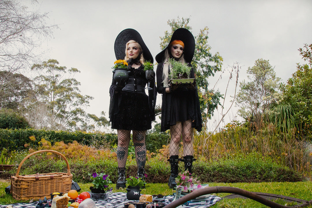 Photograph of 2 people dressed in black gothic witchy clothing with black platform boots. They are both holding plants in pots and are standing in a garden with a picnic displayed at their feet.