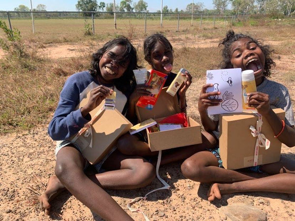 Photograph of three young girls sitting outside in the sunshine smiling at the camera. Each girl is enthusiastically showcasing their box from the Happy Boxes Project. Image supplied by Happy Boxes Project