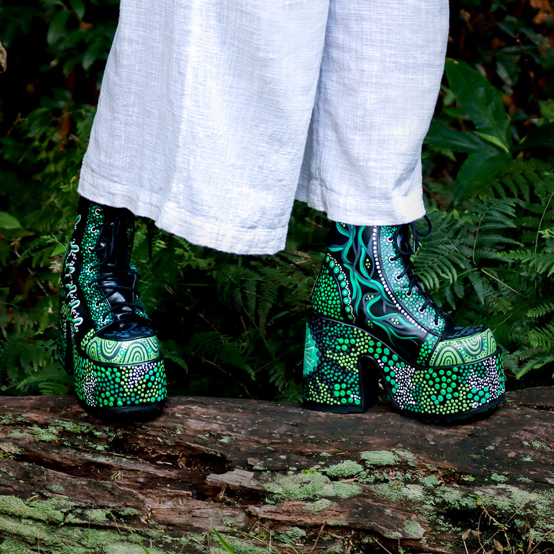 Photograph of artist Nikita Fitzpatrick wearing the hand-painted Demonia Camel-203s. The shoes are decorated with greens and whites, with the imagery designed to reflect the community of Tragic Beautiful, native bushlands, and the Traditional Custodians of the land Tragic Beautiful operates on;  the Yuggera, Turrbal, Yugarabul, Jagera and Yugambeh peoples