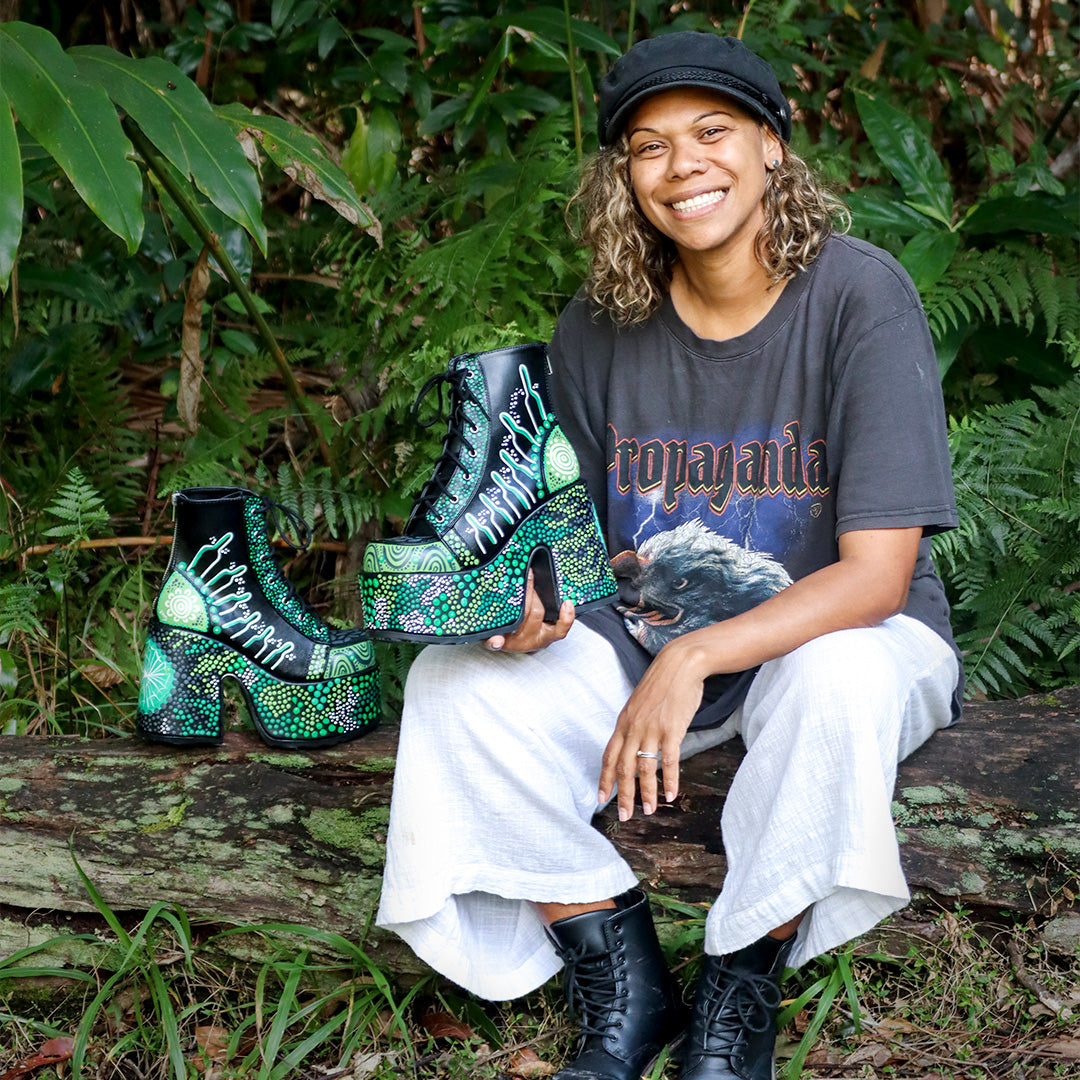 Photograph of artist Nikita Fitzpatrick displaying the hand-painted Demonia Camel-203s. Nikita is sitting outside on a fallen log with dense bushland behind her. She is wearing a dark grey t-shirt, white linen pants, and black boots. She is smiling happily at the camera.