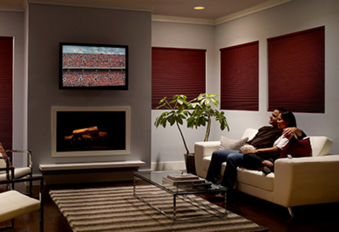 A modern living room with windows covered by red shades. There is a television mounted on the wall above a fireplace with a man and woman watching it together. The couch is beige.