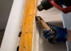 Close up of hands, one is holding a heat gun and another a scraper.  Both are removing paint off door trim.