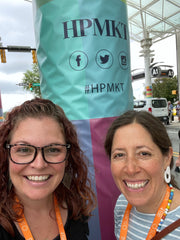 Photo of me and Marissa in front of a pillar with the HPMKT logo on it.