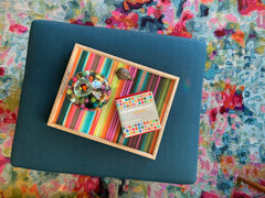 View from above of a teal ottoman with a rainbow tray.  Ottoman is sitting on a very bright and colorful rug