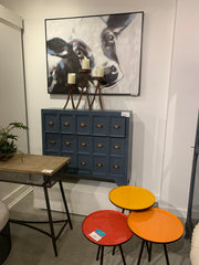 Piece of furniture with a lot of drawers, a painting of a large cow above it and 3 colorful accent tables in red, orange and yellow in the bottom right corner.