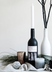 Black and white painted wine bottles, some natural branches, a pine cone and candle all set against a white backdrop