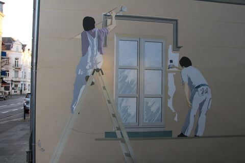 Outdoor mural of 2 people painting a window on the side of a building.