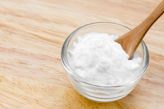 Glass bowl of baking soda with a wooden spoon in the bowl and they are sitting on a wood table.