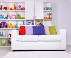 White sofa with multicolored bright accent pillows, bookcase behind sofa with a collection of rainbow items in each square section.