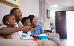 A black family with one boy and one girl child are sitting on the sofa watching television.