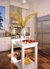 White kitchen, focus of the photo is a white mobile island in the middle of the kitchen with a vase of flowers and glasses sitting on top.