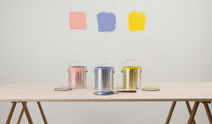 3 open paint cans on a long table, behind them on the wall are samples painted in pink, blue and yellow.