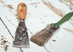 Paint scraper next to a clean paint brush on a white wooden floor