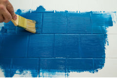 Someone using a brush to paint blue paint over a white wall tile.