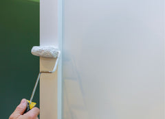 Hand with a small paint roller painting trim on a white wall with a green wall in the background
