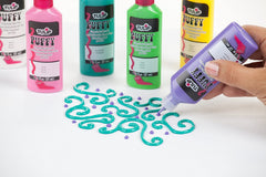 Tubes of Puffy Paint and a hand painting a curly design with a teal puffy paint.