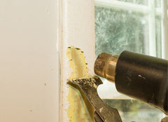 Heat gun and scrapper being used to pull paint off the trim of a window.
