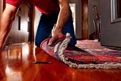 Muscular man on hands and knees applying rug anchor to a corner of a rug.