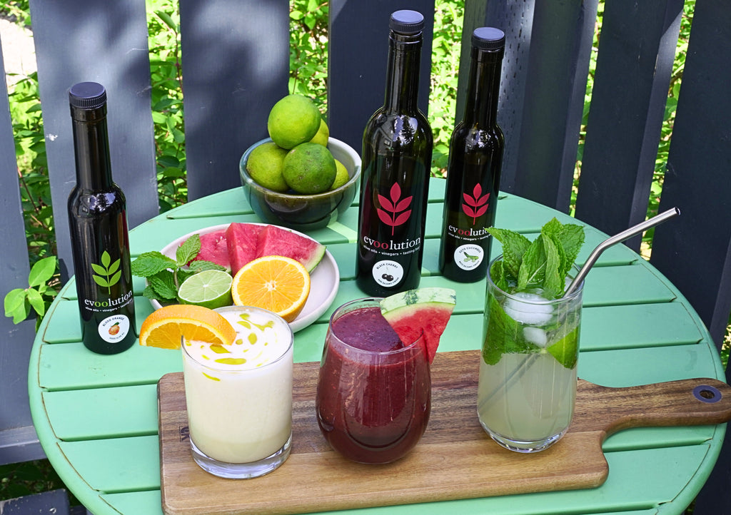 A green metal table holds a wooden serving board with three cocktails, accompanied by fresh fruits and herbs and three bottles of Evoolution oil and vinegar