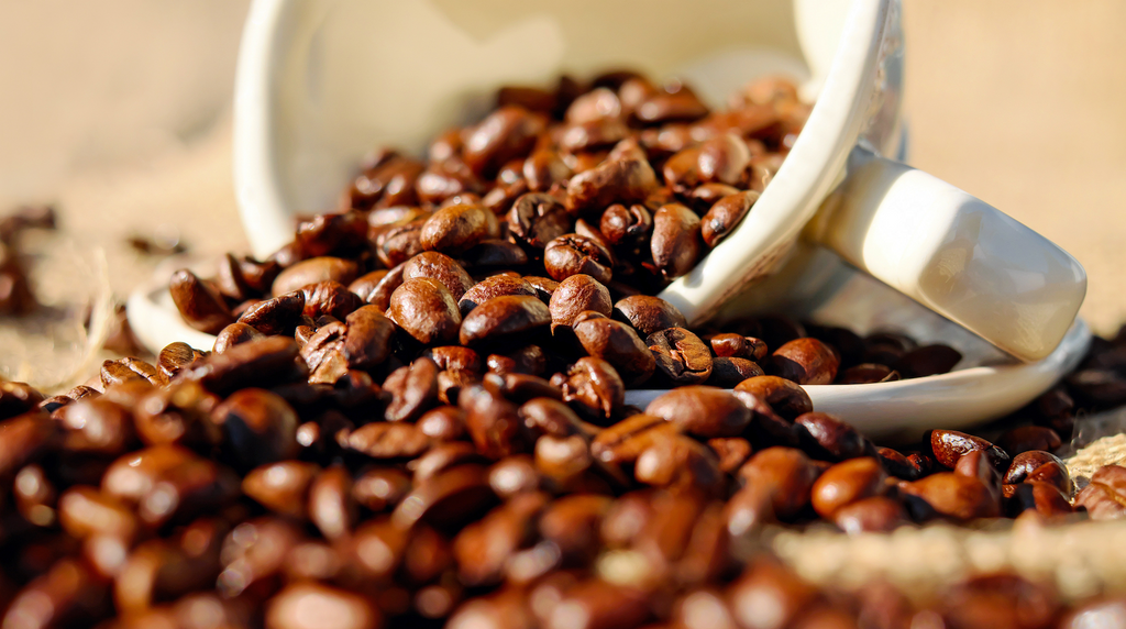 A white mug full of coffee beans spills its contents on to a table.