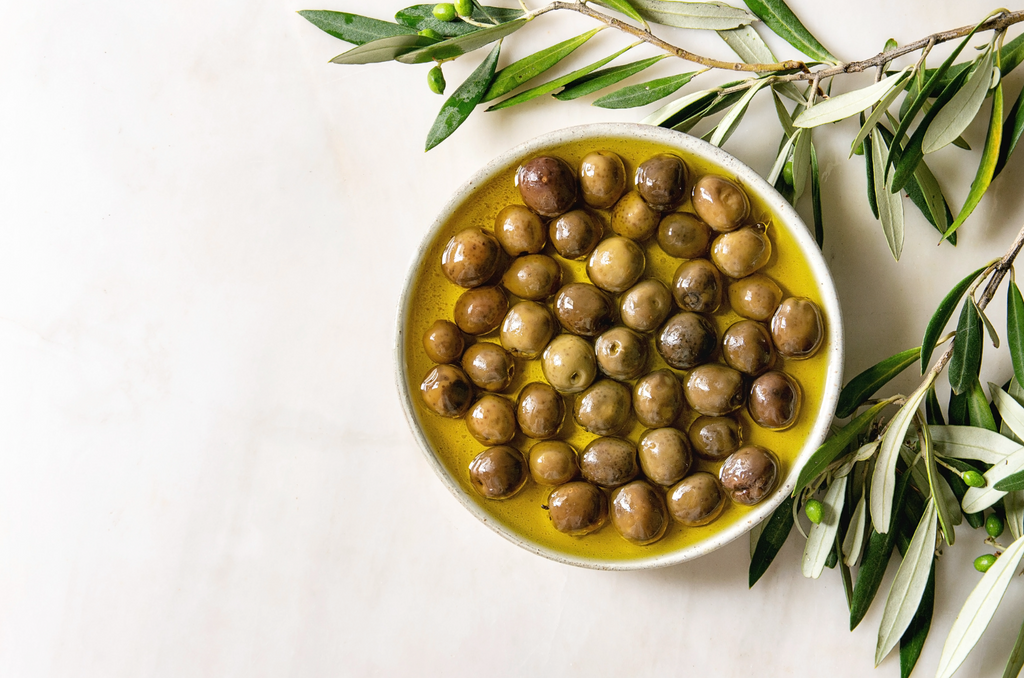Bowl of olives marinated in EVOO surrounded by olive leaves.