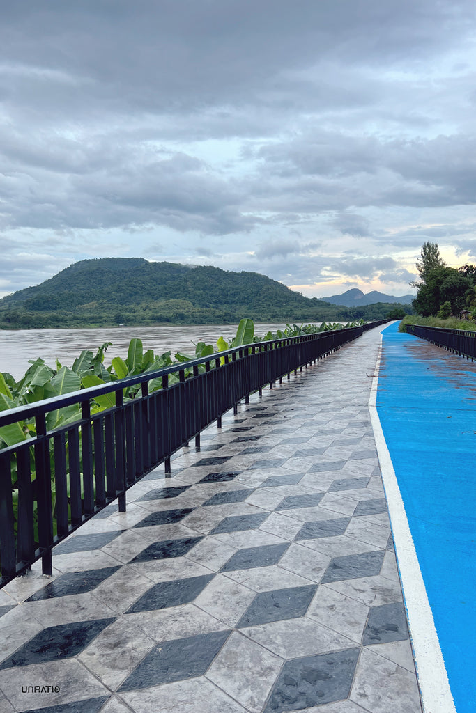 Scenic Chiang Khan bike path with geometric tiles alongside the lush Mekong River under a dramatic cloudy sky, encapsulating the serene beauty of northeastern Thailand.