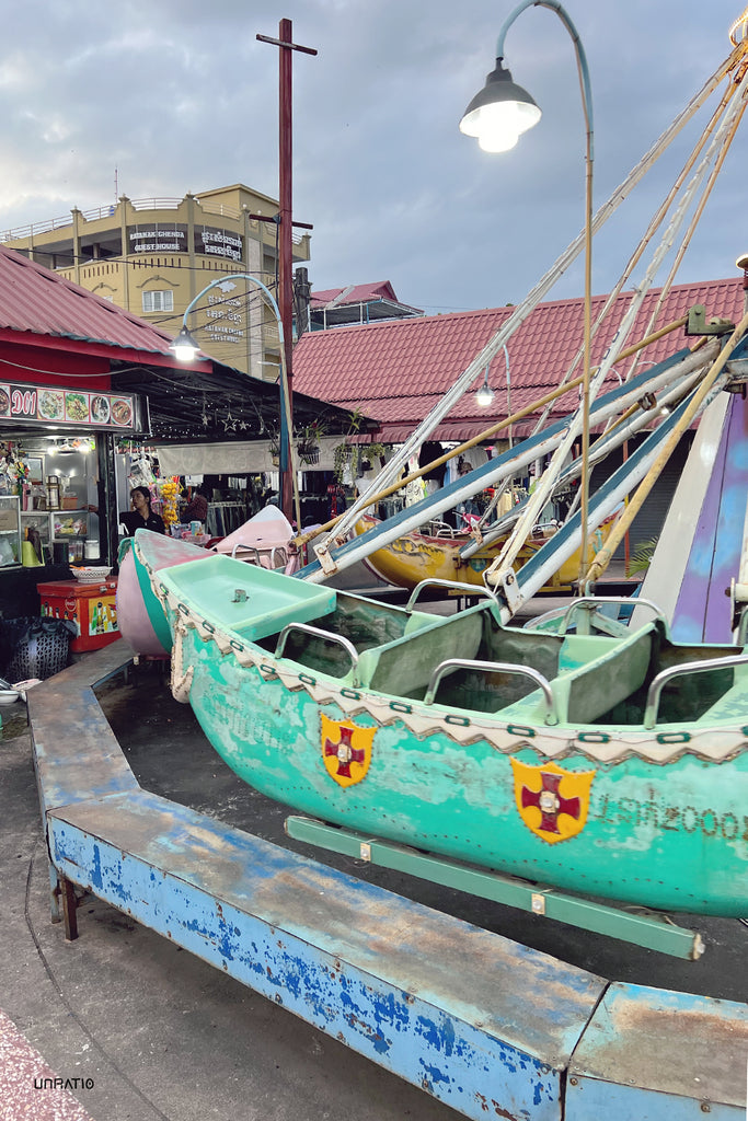 Early evening at Kampot market with a view of a whimsical carousel featuring turquoise boats, ready for nighttime riders, amidst the bustling local vendor stalls.