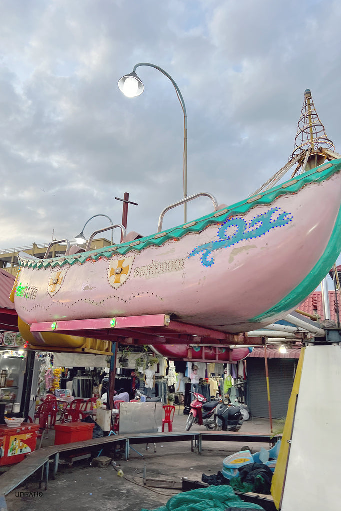 Kampot evening fair with a close-up of a nostalgic carousel boat in pastel pink, lit by delicate lights under a cloudy sky, with market life stirring in the background.