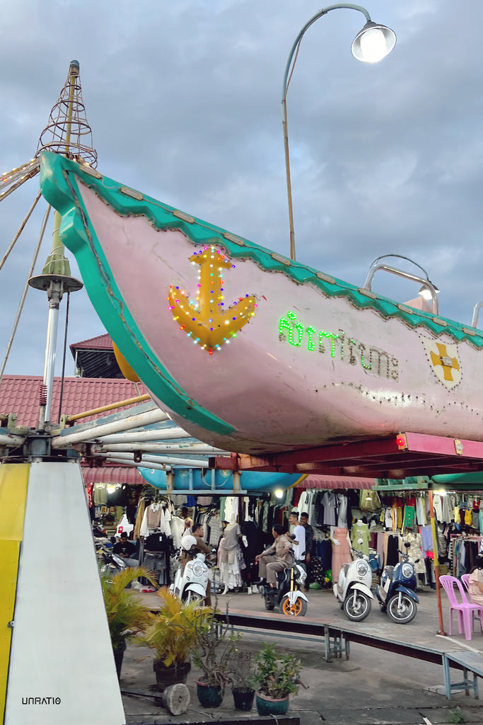 Evening view of a pink-hued carousel boat with vibrant light decorations in Kampot, as local life buzzes in the market background with shoppers and scooters.