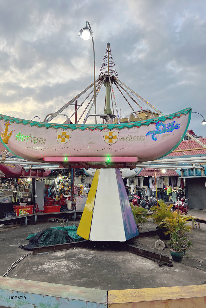 Twilight at Kampot fair with a vintage carousel ride, pastel-painted boats adorned with lights, against a backdrop of a soft sky and local market stalls.