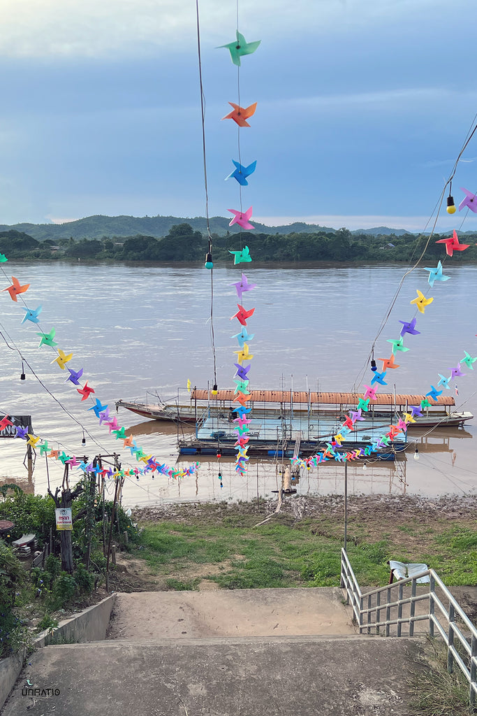 Colorful pinwheel decorations strung above the tranquil Mekong River with traditional Thai boats docked along the bank, reflecting Chiang Khan's vibrant community spirit.