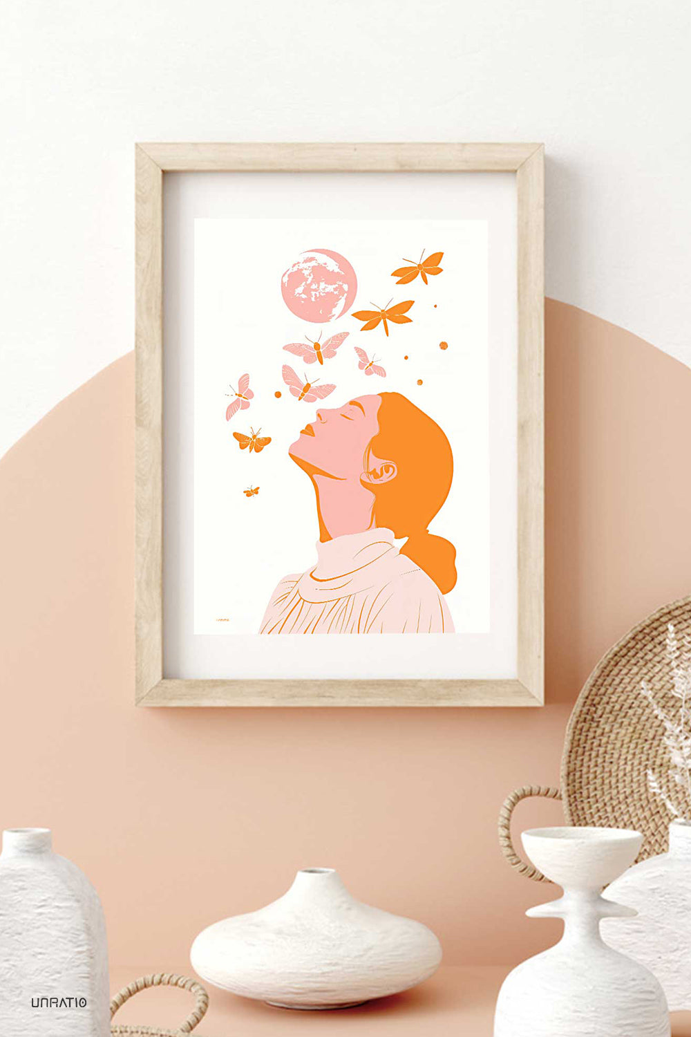 Interior decor scene with a framed print of a woman and a pink moon with moths, placed on a wall above modern white vases.