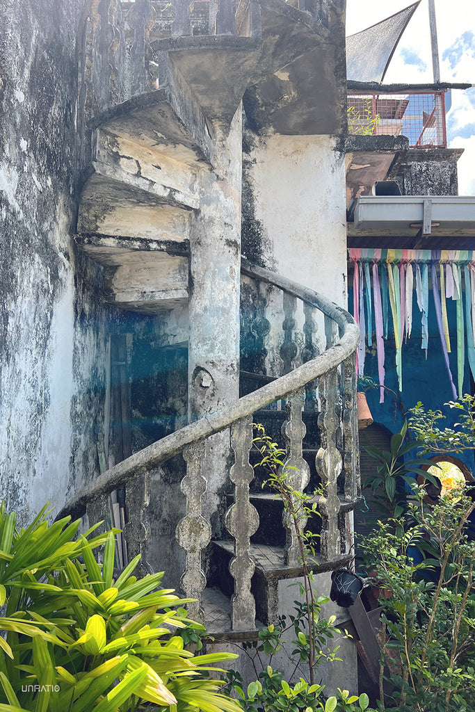 Antique spiral staircase with intricate balusters in a lush Melaka courtyard, exuding old-world charm.