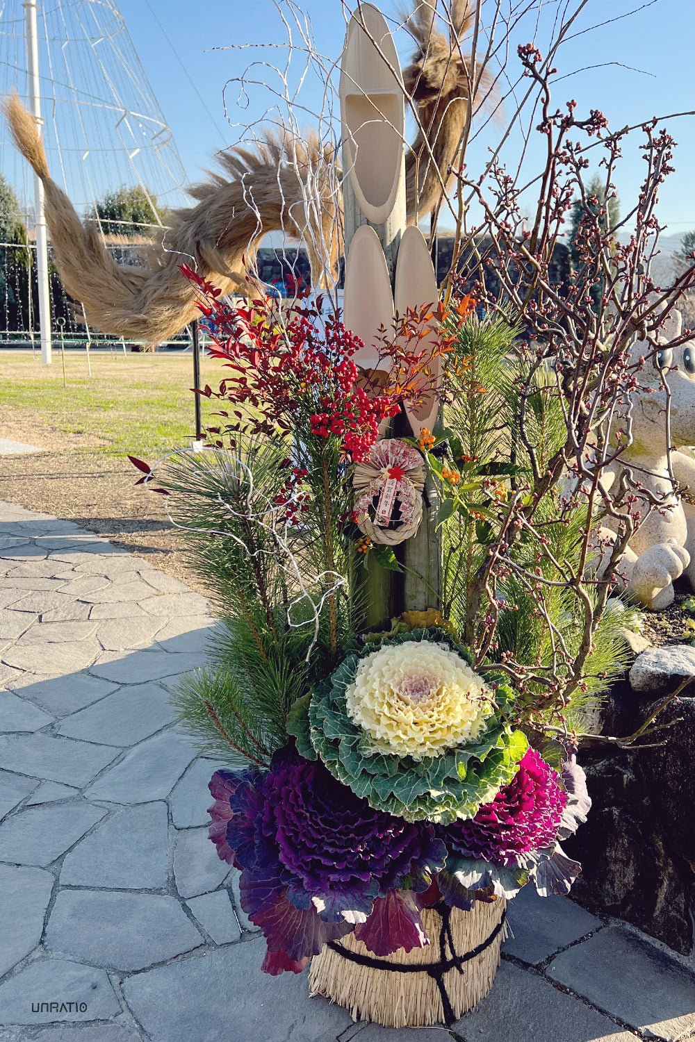 Elaborate floral arrangement featuring ornamental cabbages, pine branches, and bright berries, set against a park with artistic sculptures.