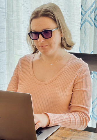 A woman with shoulder-length hair wearing sunglasses and a pale pink long-sleeved top is working on a laptop. She appears focused on the screen, with the laptop placed on a table in a room with natural light coming through a window with patterned curtains in the background.