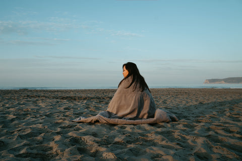 Woman wrapped in the Gobi Heat Zen blanket sitting on the beach