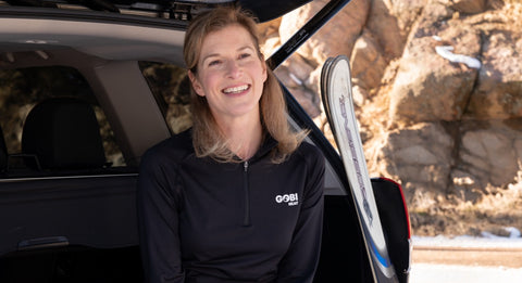 Woman sitting in an open trunk of a car with ski gear wearing the Gobi Heat Basecamp base layer shirt