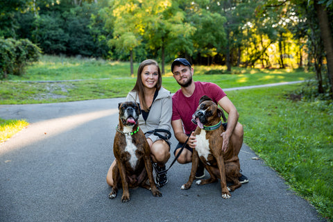 Man and woman sit in driveway with their two Boxer dogs.