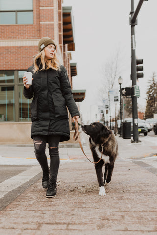 Blonde woman in black jacket and pants walks big black dog outside using Mighty Paw Leash.