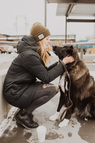 Blonde woman in black coat and pants gives big black dog a kiss.