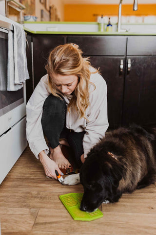 Woman is on floor cutting her dog's nails with Mighty Paw Nail Clippers while dog licks Mighty Paw Lick Pad.