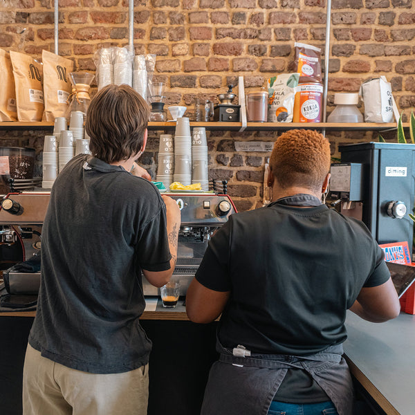 Two women working in a coffee shop with their backs to the camera facing a coffee machine