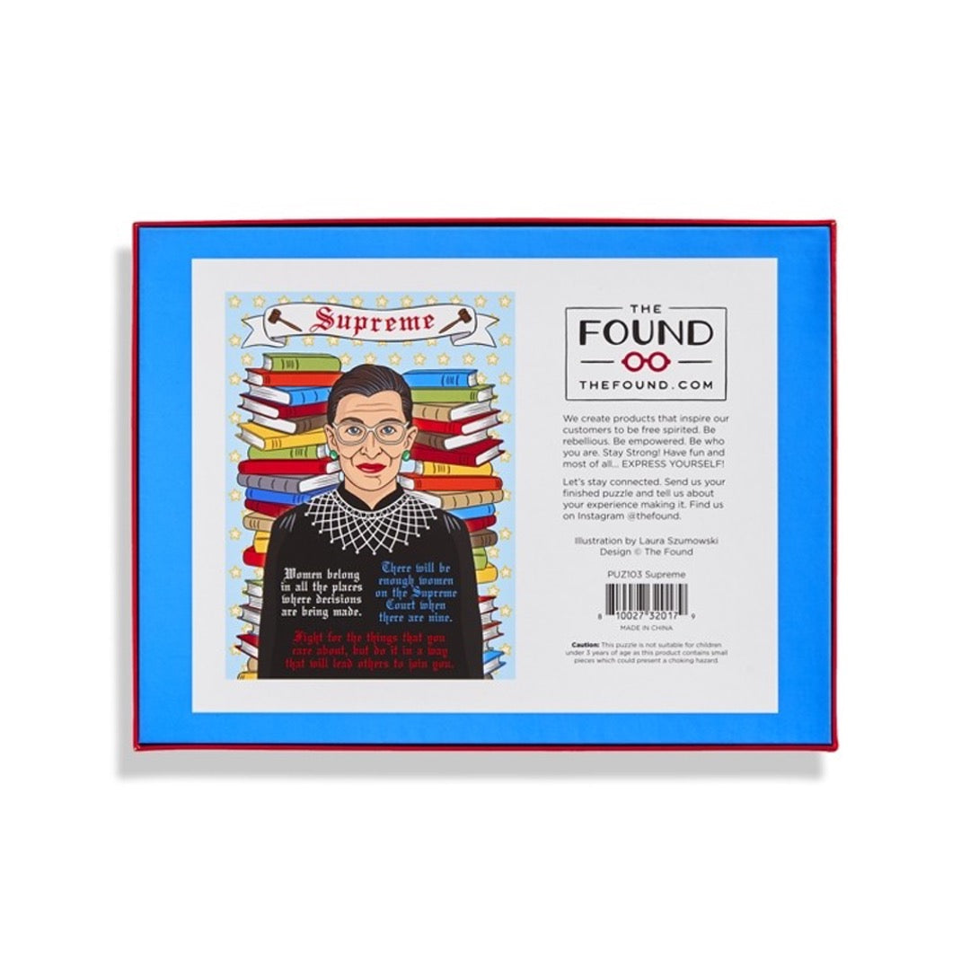 Back 500 piece puzzle box of Ruth Bader Ginsburg in front of stack of books with some of her famous quotes. Banner on top that says Supreme and 2 gavels on either side