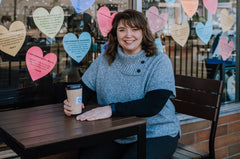Christine is sitting at a table with the window of Stonehouse Teas covered in hearts behind her