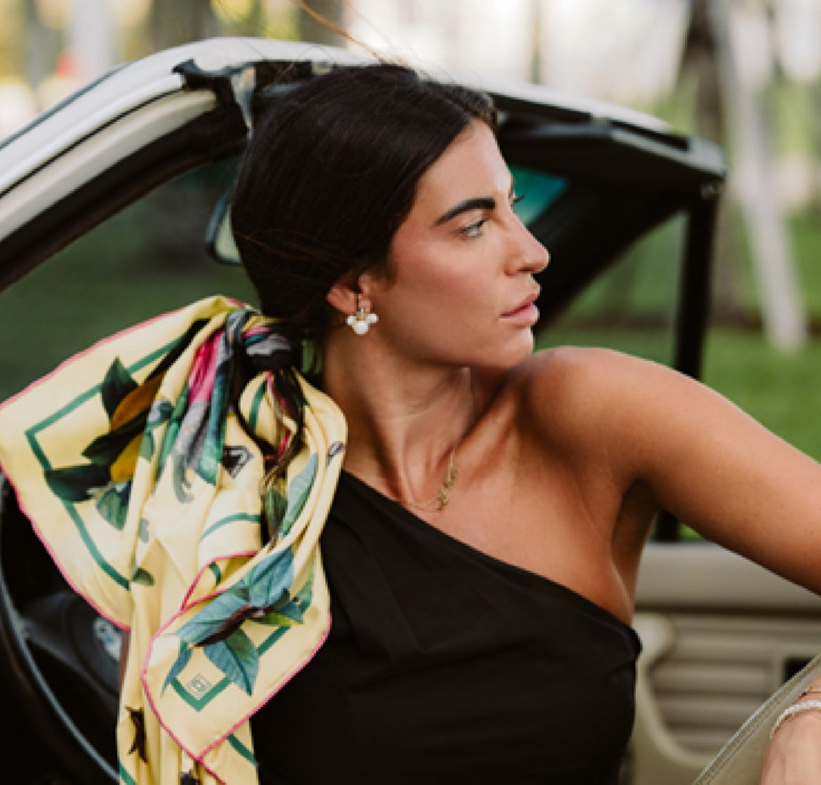 Brunette with off the shoulder top wears scarf in her hair while sitting in convertible.