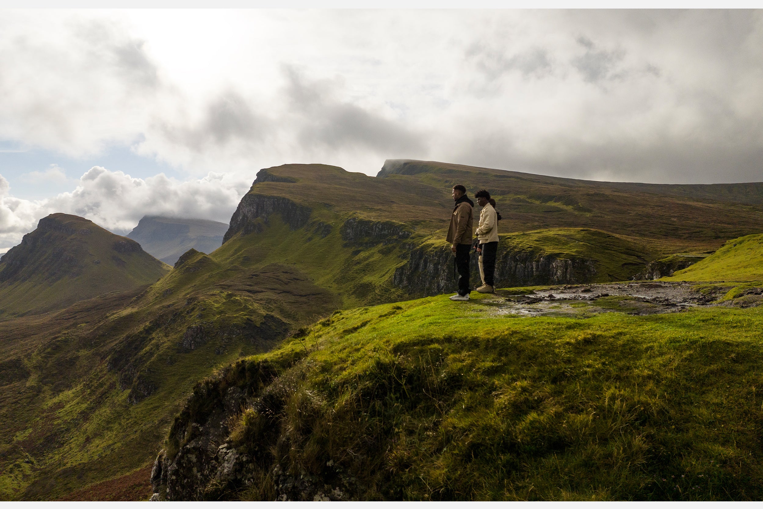 Three male models in black cargo pants and jackets stand atop the Isle of Skye.