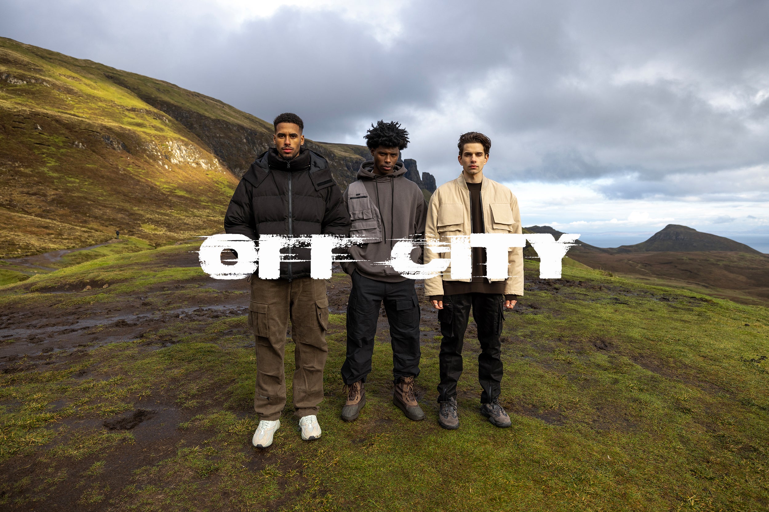 Three male models in black cargo pants and jackets stand atop the Isle of Skye.