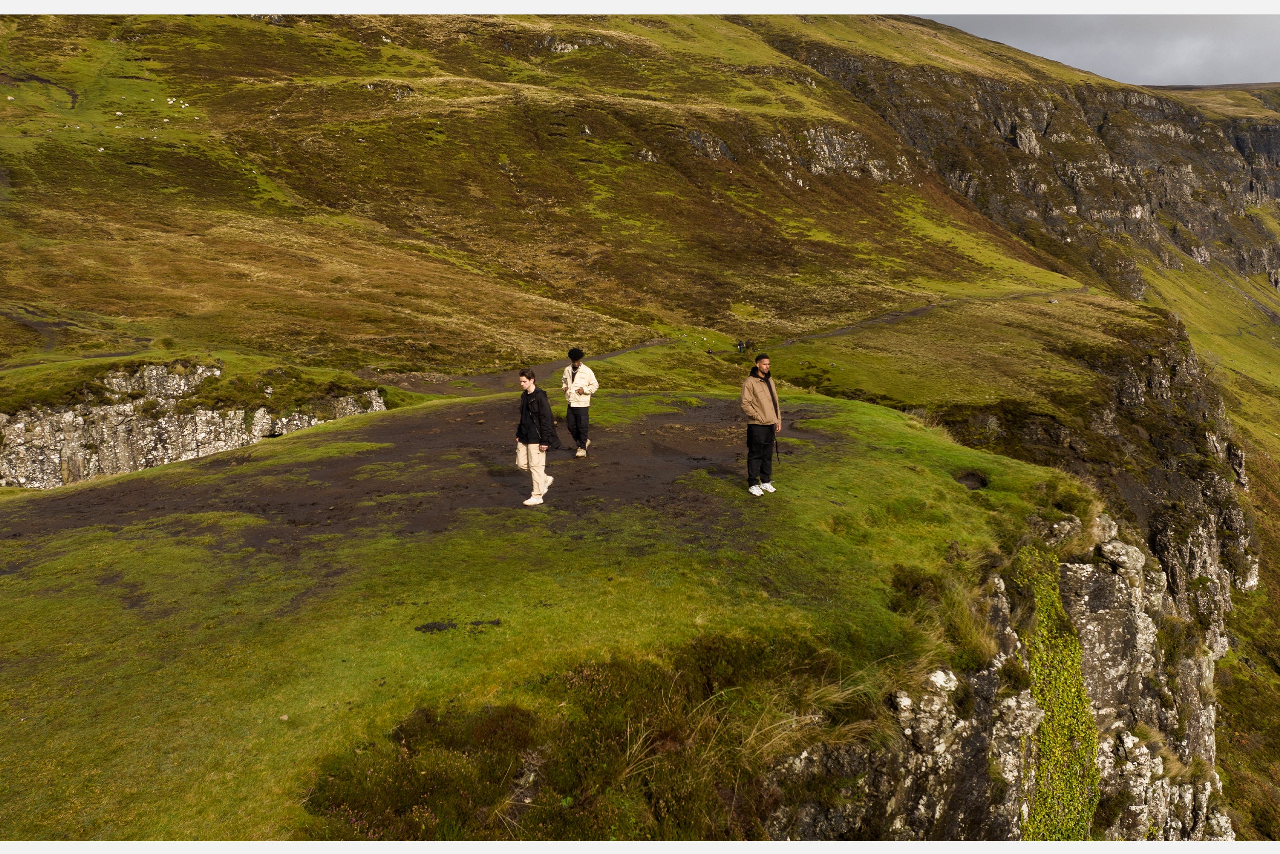 Three male models in black cargo pants and jackets stand atop the Isle of Skye.