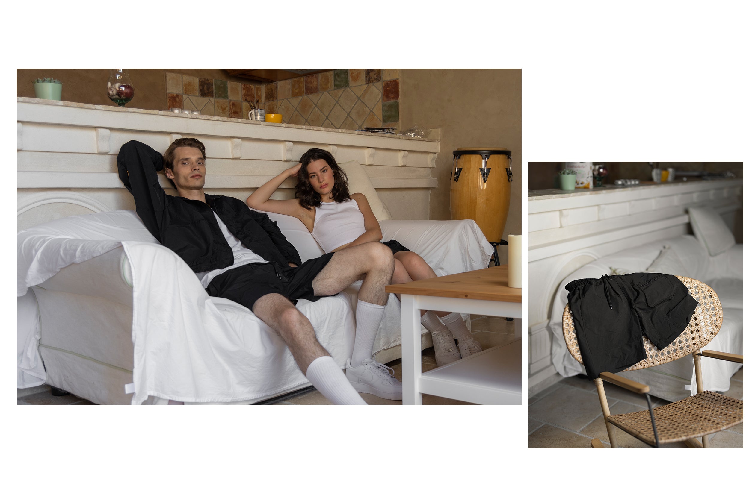 A male model wearing a black zip-up jacket and black cargo shorts is sitting on a sofa next to a female model wearing a white tank top and black cargo shorts.